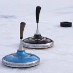 Zwei Eisstöcke liegen auf einer Eisfläche; dahinter liegt ein schwarzer Puck.