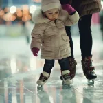 Kleines Kind in Winterkleidung läuft auf Schlittschuhen und hält sich an einer erwachsenen Begleitperson fest.