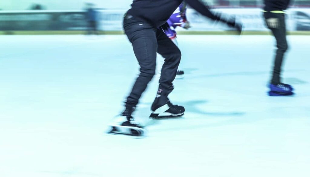 Einzelpersonen laufen auf einer Eisbahn in Deggendorf Schlittschuh. Ein Schlittschuhläufer im Vordergrund ist mitten im Schritt aufgenommen, er trägt dunkle Hosen und ein dunkles Oberteil. Das Bild hat einen Bewegungsunschärfeeffekt, der die Bewegung und Geschwindigkeit betont. Der Hintergrund besteht aus einer Indoor-Eisbahnkulisse für einen öffentlichen Eislauf.