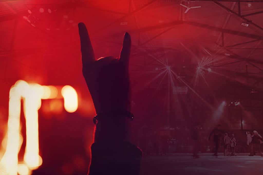 Vor einem rot beleuchteten Konzert- oder Veranstaltungsort wird eine Handsilhouette erhoben, die eine „Rock on“-Geste macht, was an die Energie bei einem Eislauf-Rockkonzert erinnert. Im Hintergrund sorgen verschwommene Figuren und Bühnenlichter für eine lebendige Atmosphäre.