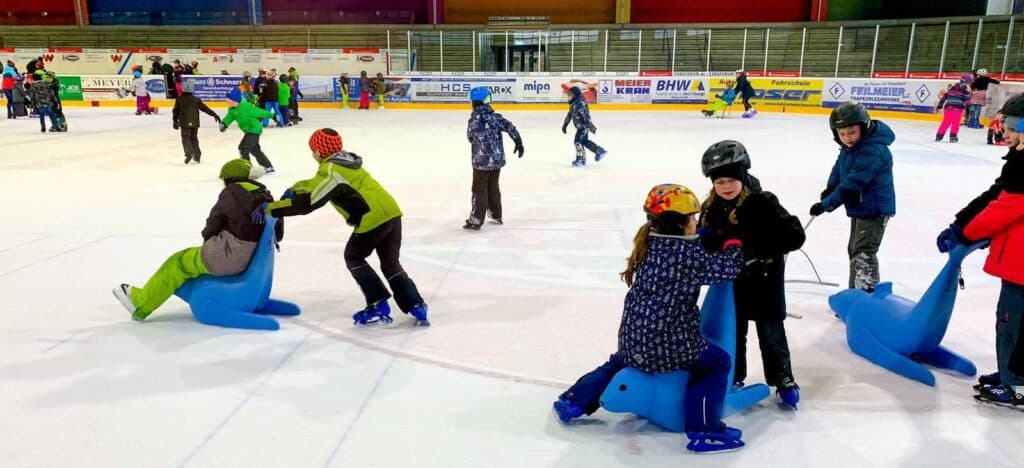 Kinder, die in einer Eishalle Schlittschuh laufen. Einige benutzen blaue, siegelförmige Eislaufhilfen, während andere Schlittschuh laufen oder herumstehen. Im Hintergrund sind weitere Schlittschuhläufer und die Absperrung der Eisbahn mit Werbung für den Schul-Eislauf Deggendorf zu sehen.